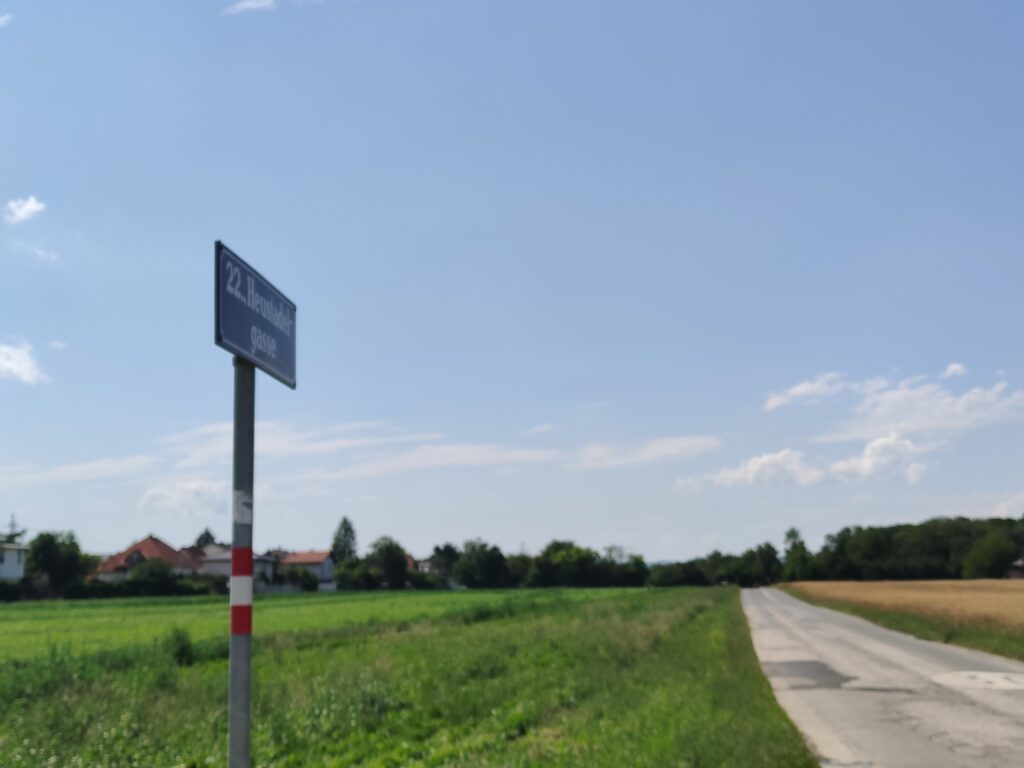 Straßenname am Rande der Heustadelgasse. Die Straße ist alt-betoniert. Links und rechts ovn der Straße sind grüne Wiesen und Weizenfelder. Im Hintergrund eine Siedlung umgeben von einem Wald. Alles unter schönem Sommerhimmel.