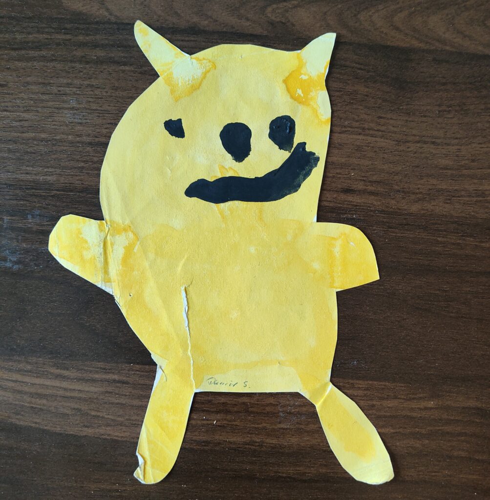 Papier-Ausschnitt eines gelben Tieres, dass aussieht wie Pikachu, aber ohne Schwarz. Mit dicker schwarzer Tusche ist ein krummes Gesicht draufgemalt.